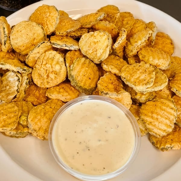 A white plate piled high with crispy fried pickle chips and a side of creamy dipping sauce.