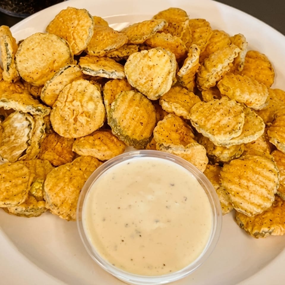A white plate piled high with crispy fried pickle chips and a side of creamy dipping sauce.