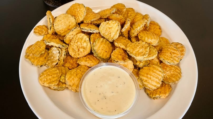 A white plate piled high with crispy fried pickle chips and a side of creamy dipping sauce.