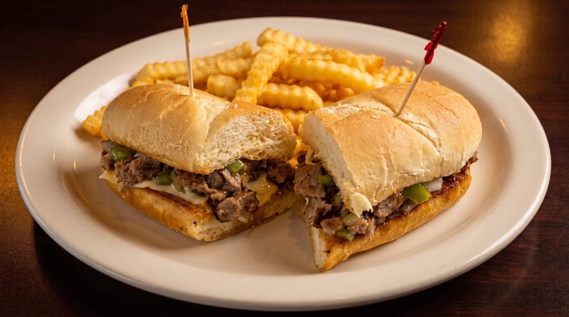 A cheesesteak sandwich, cut in half, with crinkle-cut fries on a white plate.