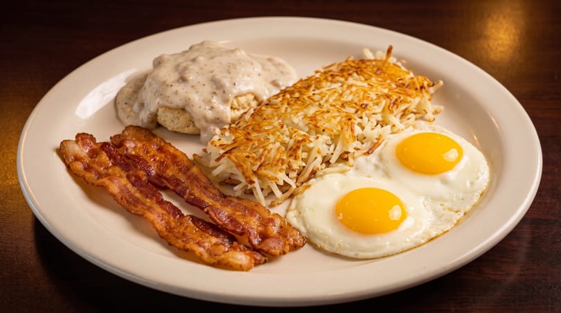 A breakfast plate with biscuits and gravy, hash browns, two fried eggs, and bacon.