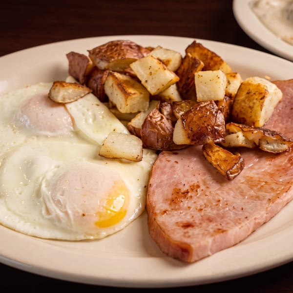 Breakfast plate with two fried eggs, crispy home fries, and a slice of grilled ham.