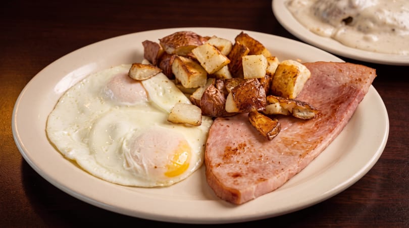 Breakfast plate with two fried eggs, crispy home fries, and a slice of grilled ham.