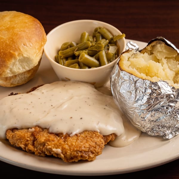 Chicken fried steak with gravy, baked potato, green beans, and dinner rolls on a white plate.