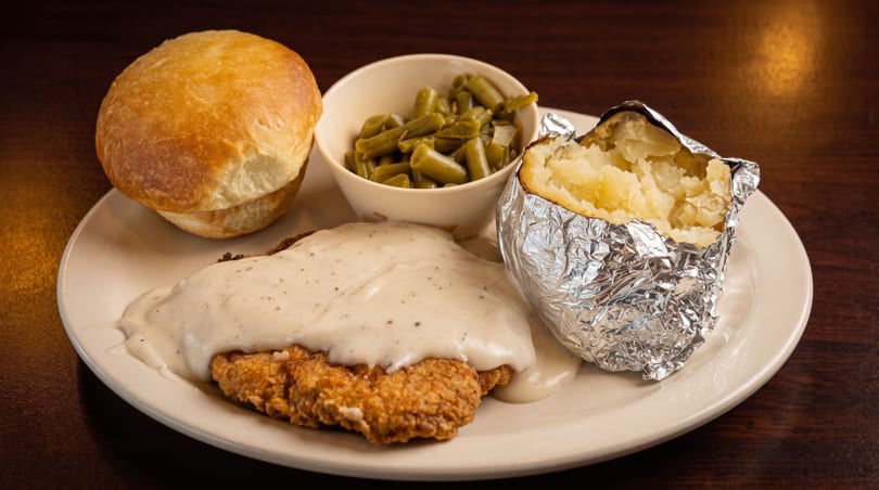 Chicken fried steak with gravy, baked potato, green beans, and dinner rolls on a white plate.