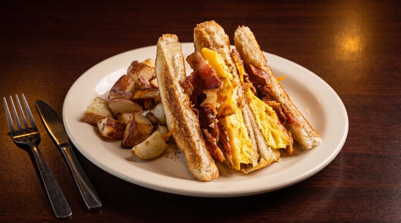 Breakfast sandwich with scrambled eggs, bacon, and cheese, served with roasted potatoes on a white plate.