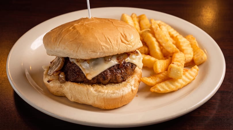 Cheeseburger with melted cheese, caramelized onions, and mushrooms, served with crinkle-cut fries on a white plate.