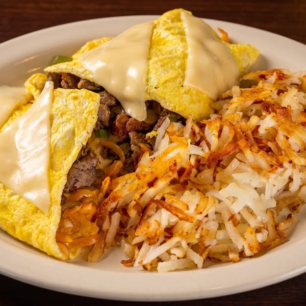 Cheesesteak omelet with melted cheese and hash browns on a white plate.