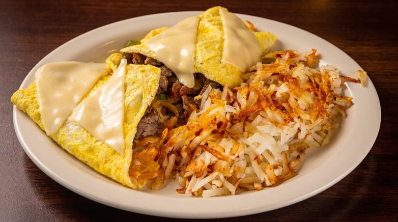 Cheesesteak omelet with melted cheese and hash browns on a white plate.