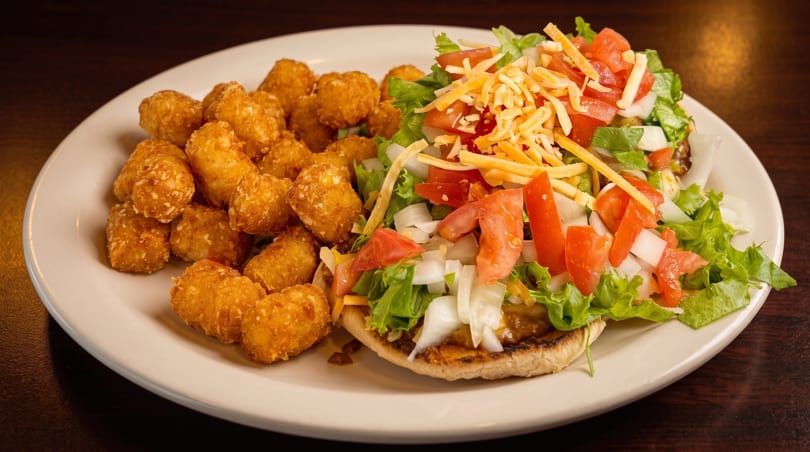 A plate of golden tater tots next to an open-faced burger topped with lettuce, tomato, onion, and cheese.