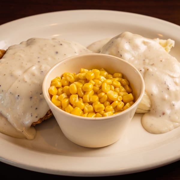 Chicken fried steak with gravy, mashed potatoes, and a side of corn on a white plate.