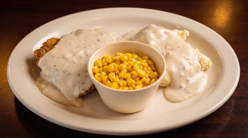 Chicken fried steak with gravy, mashed potatoes, and a side of corn on a white plate.