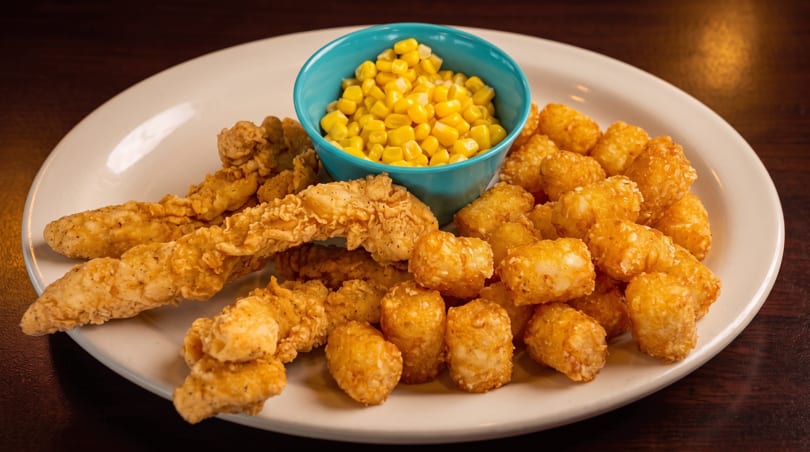 Crispy chicken tenders, golden tater tots, and a teal bowl of corn on a white plate.