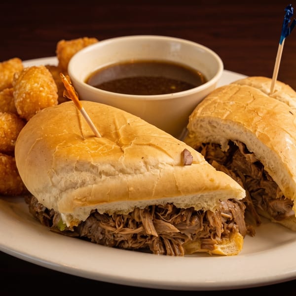Two French dip sandwiches with tater tots and a side of au jus on a white plate.