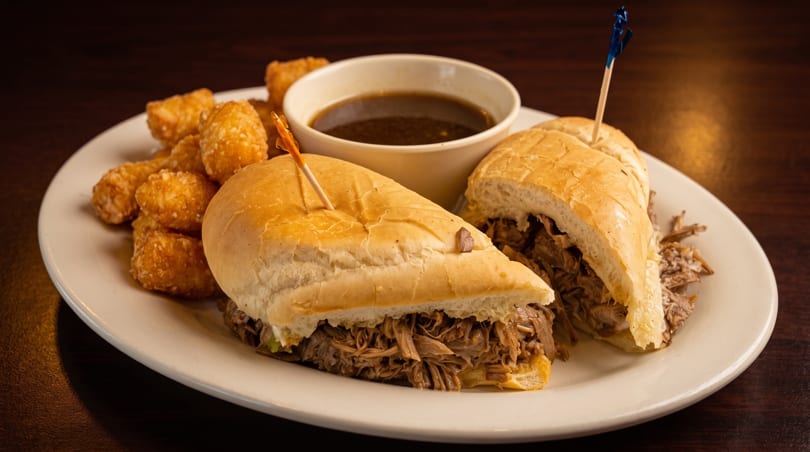 Two French dip sandwiches with tater tots and a side of au jus on a white plate.