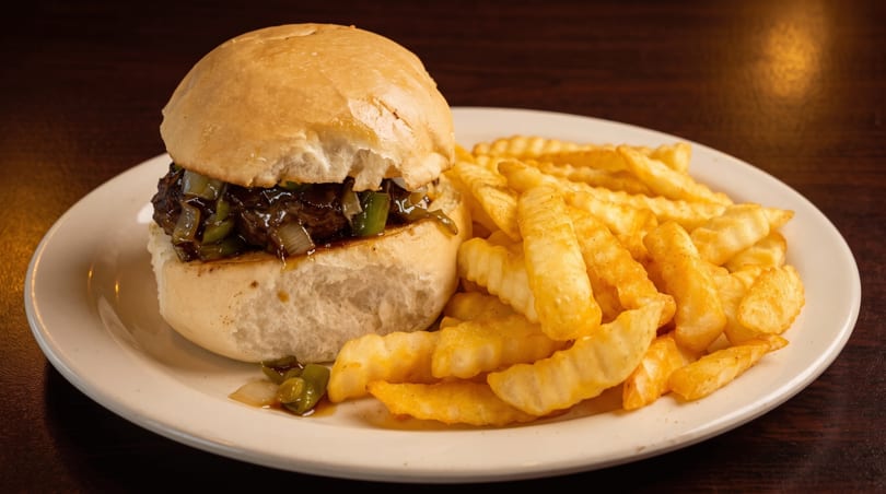 A slider with dark meat, green peppers, and onions, served with crinkle-cut fries on a white plate.