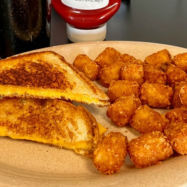 Grilled cheese sandwich halves and tater tots on a plate, with a drink and ketchup in the background.