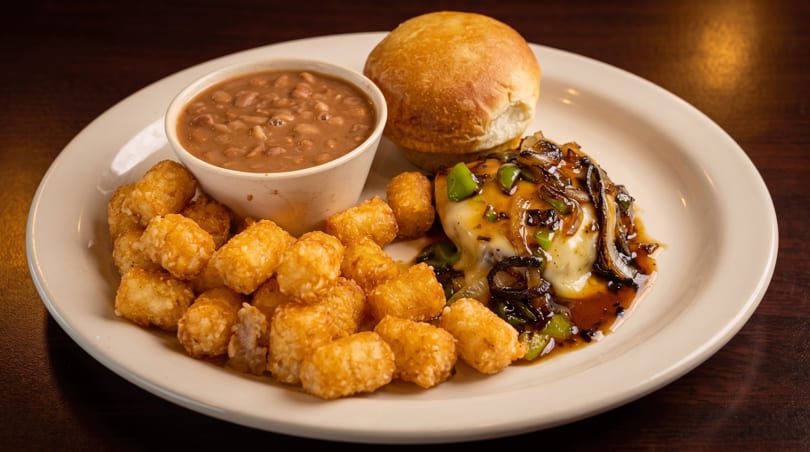 A plate with a hamburger steak, tater tots, baked beans, and a dinner roll.